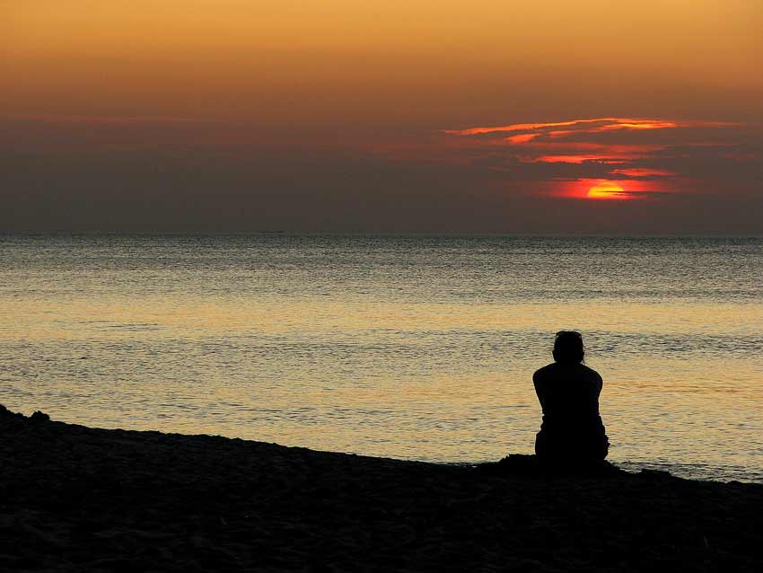 Persona sentada en la playa viendo el caer del sol. La persona está de espaldas a la cámara y se ve su silueta. 