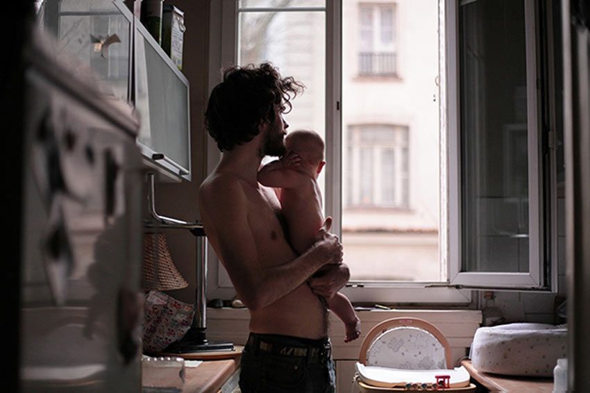 Hombre cargando a un bebé en su cocina. Ambos miran hacia la ventana.
