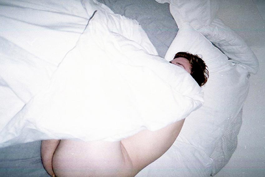 Persona acostada, desnuda con una sábana encima. Se logra ver sus gluteos y parte de su espalda.