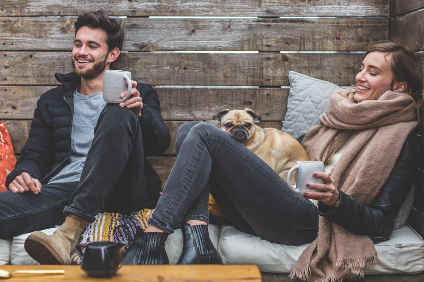 Hombre y mujer sentados riendo y tomando café. En las piernas de la mujer hay un perro.