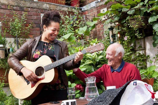 En la imagen una joven tocando guitarra a un hombre de edad avanzada en un patio