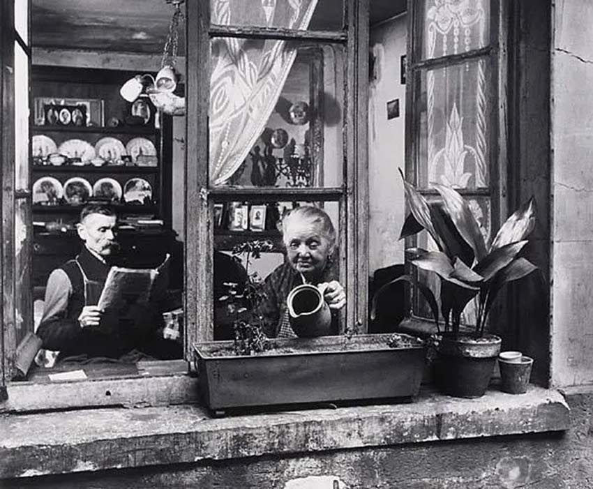 Fotografía: Robert Doisneau. Fotografía de una mujer regando plantas en una ventana. Hay un hombre leyendo el diario.