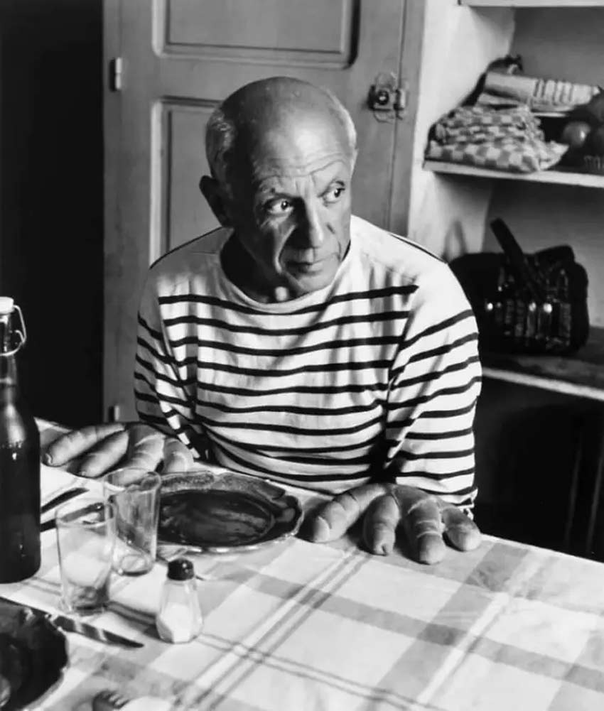 Fotografía: Robert Doisneau. Un hombre sentado mirando hacia al lado derecho, sobre una mesa unos panes.