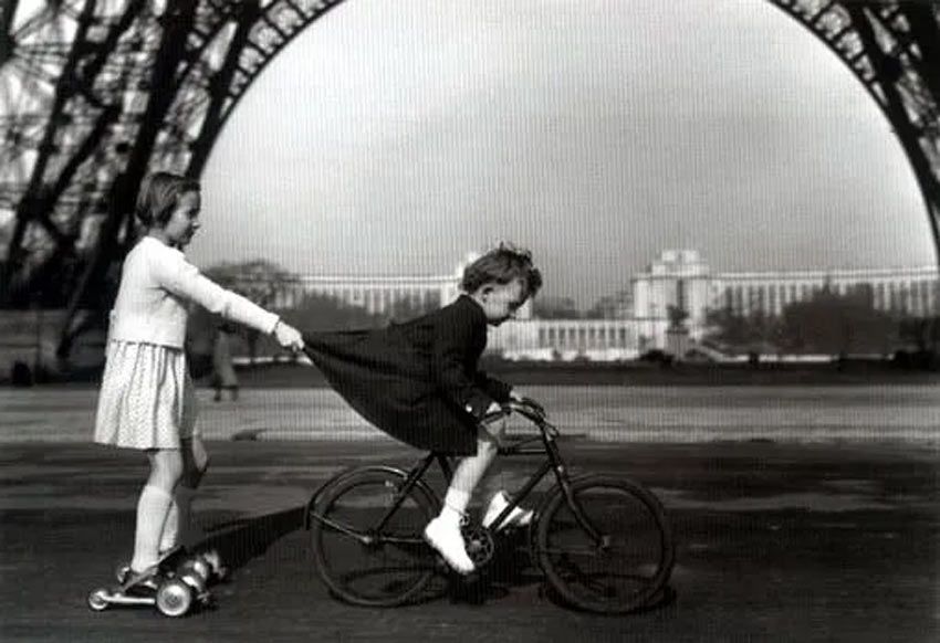 Fotografía: Robert Doisneau. Niño sobre una bicicleta y niña impulsada por el niño en sus patines.