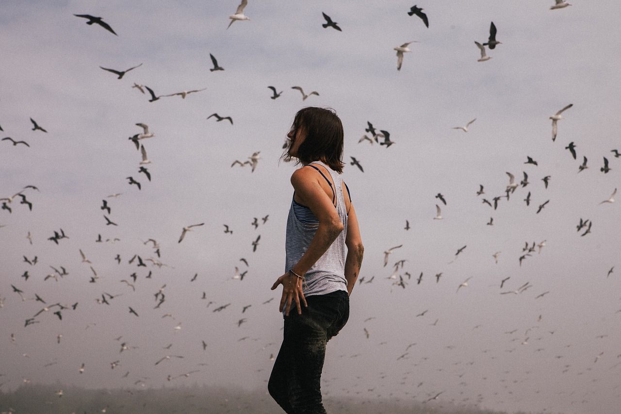 Mujer con cabello a la altura de sus hombros, de perfil a la cámara. Está parada y alrededor muchas aves costeras.